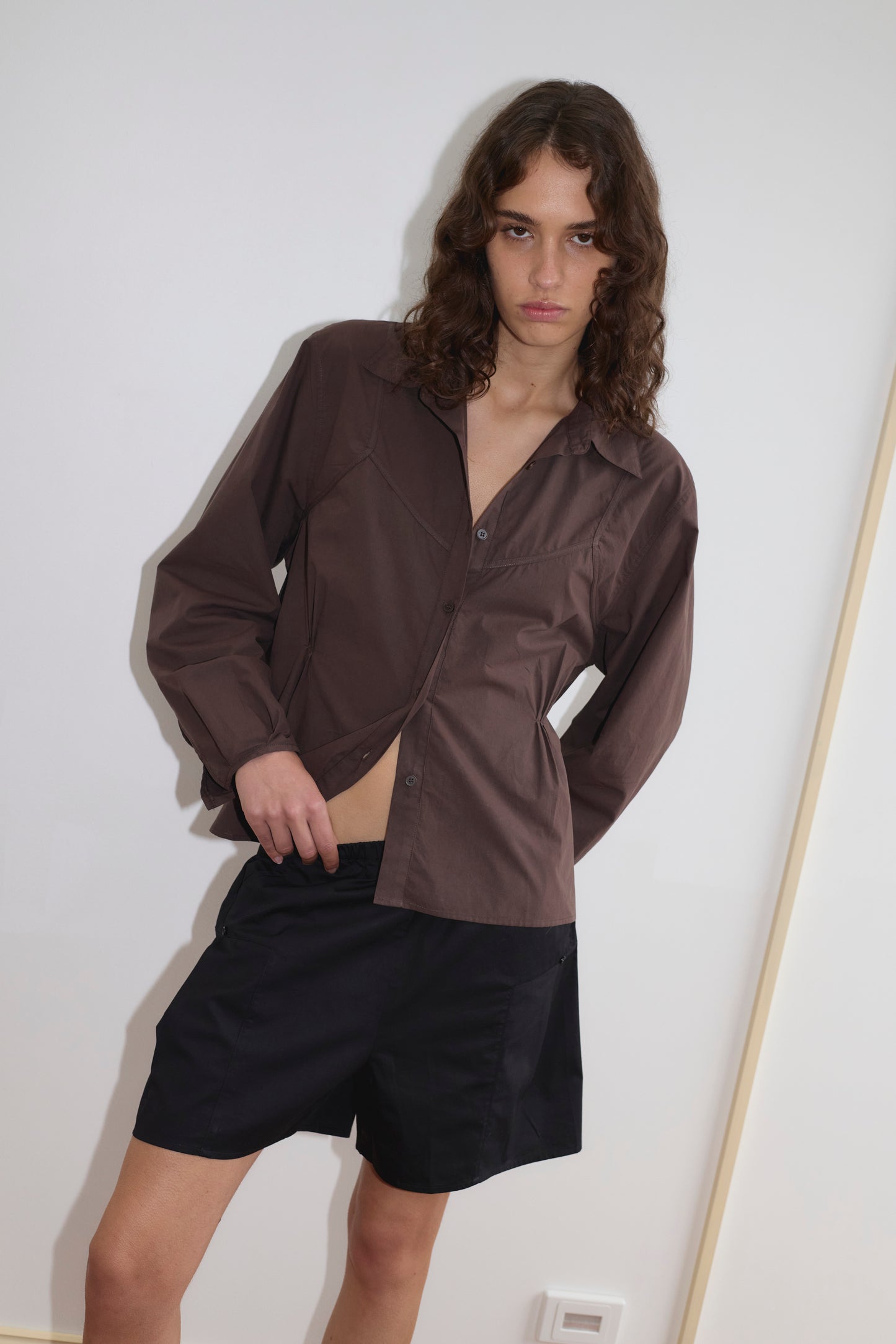 A person with long wavy hair wears a brown Deiji Studios button-up shirt and black Pleat Shorts, standing against a plain white wall with a neutral expression while adjusting the shirt.