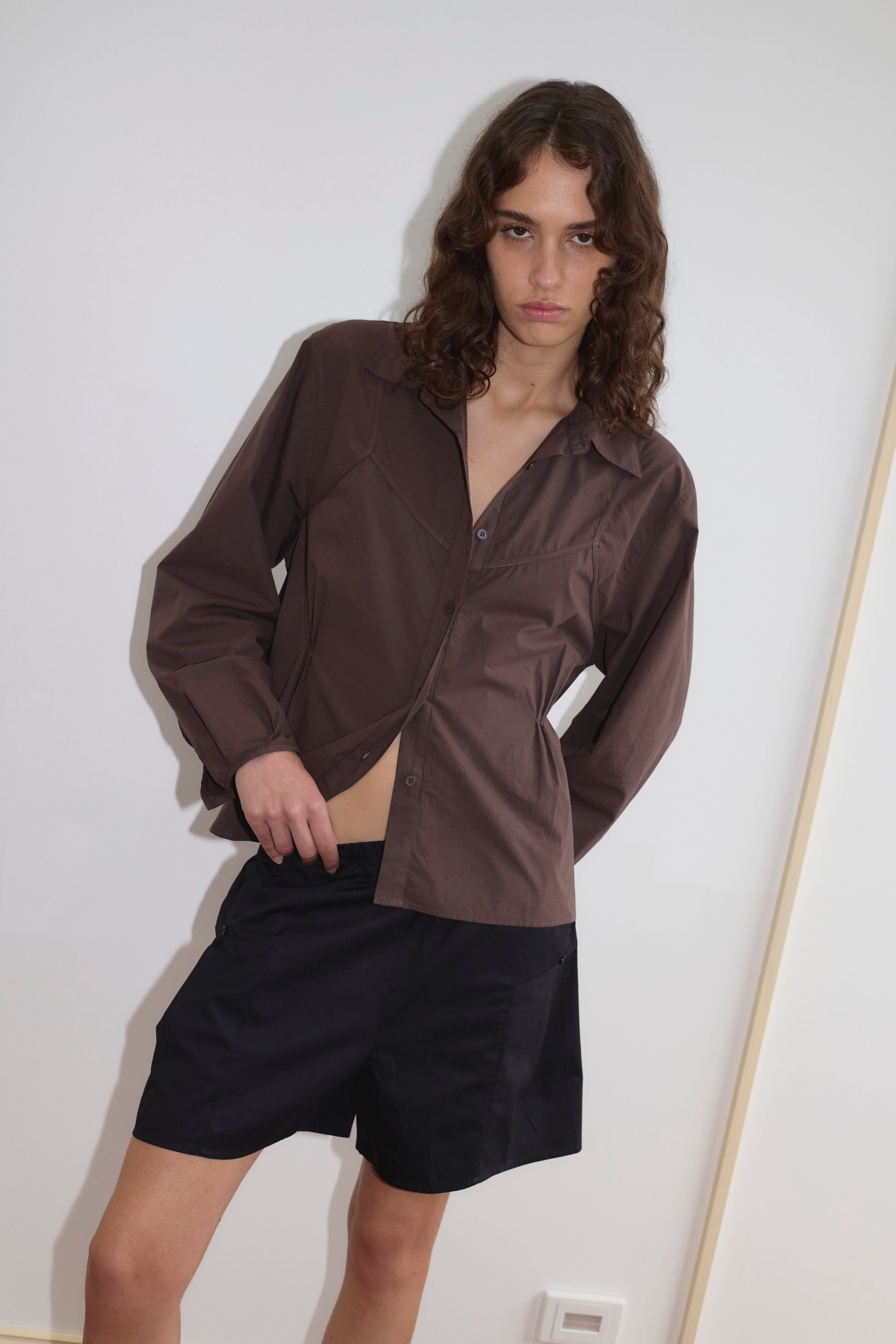 A person with long wavy hair wears a brown Deiji Studios button-up shirt and black Pleat Shorts, standing against a plain white wall with a neutral expression while adjusting the shirt.