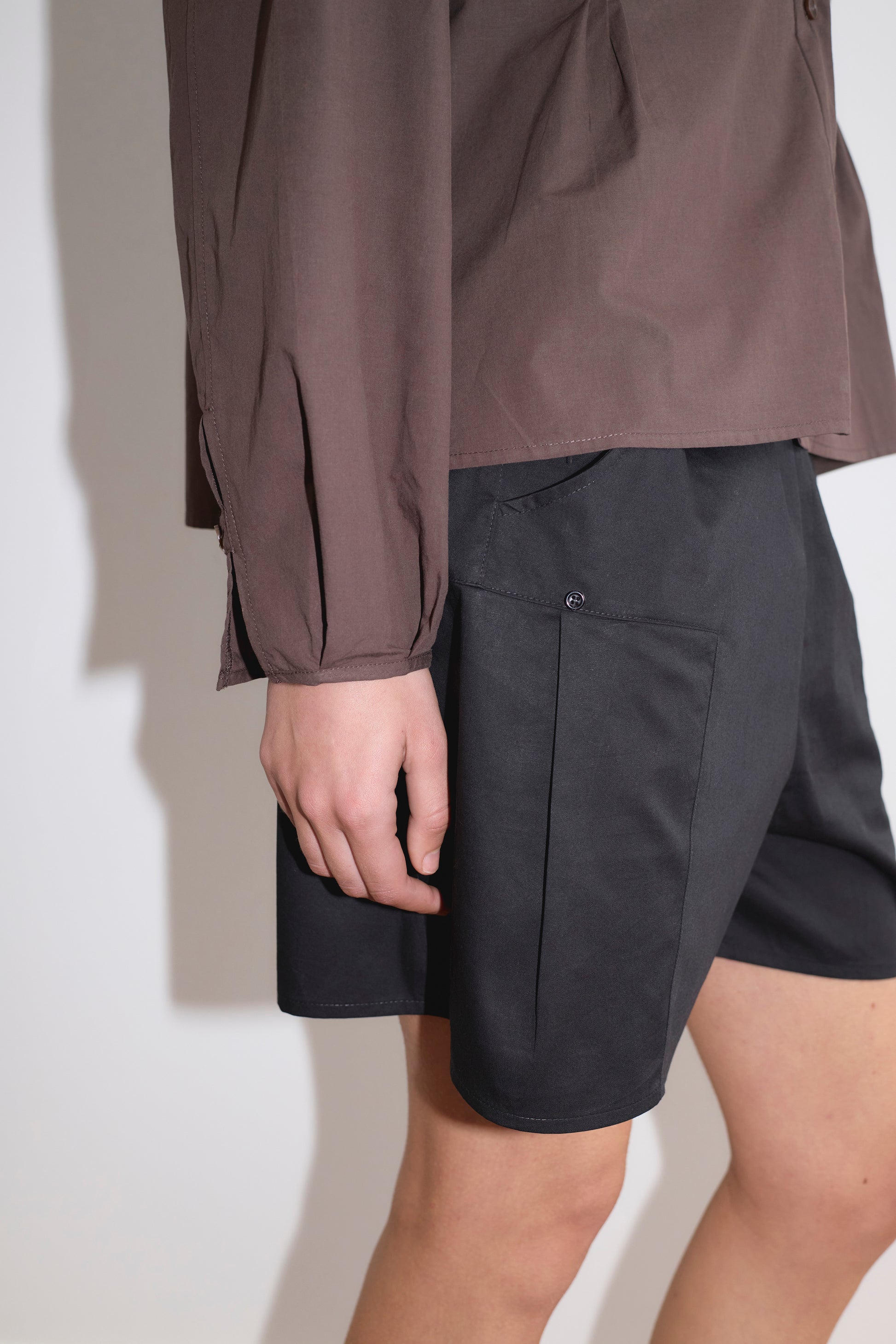 A person wearing a long-sleeved brown shirt and Deiji Studios Pleat Short in black stands against a plain white background, their left arm relaxed by their side.