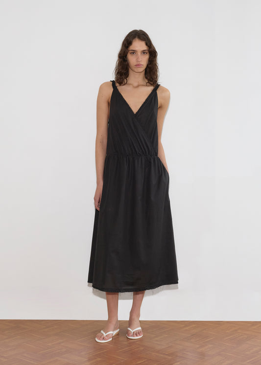 A woman with wavy brown hair wears the Crossed Dress - Black by Deiji Studios and white sandals, standing on a wooden floor against a plain white wall.