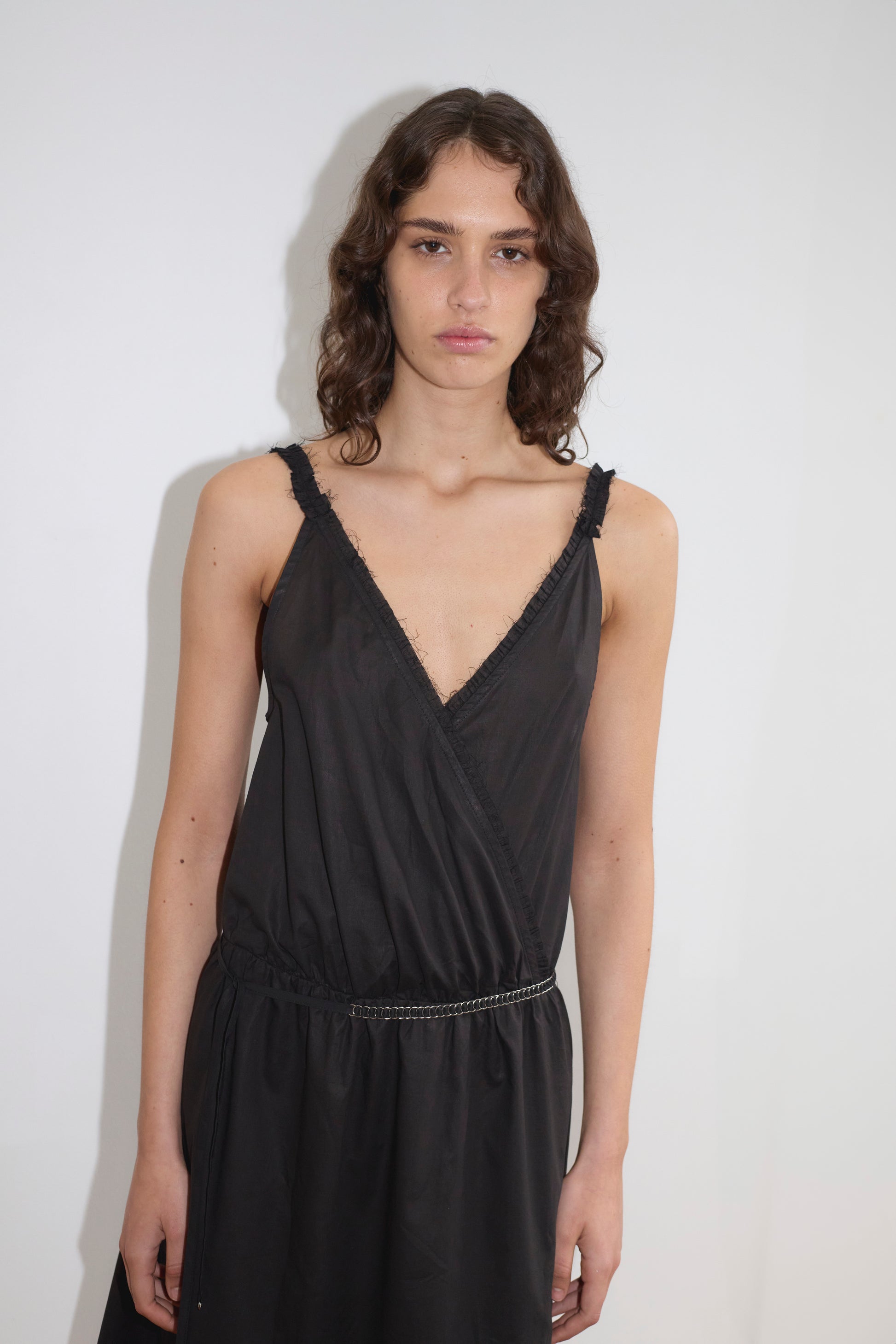 A person with wavy brown hair wears the Crossed Dress - Black by Deiji Studios, sleeveless and midi-length, standing against a plain white background and looking straight at the camera with a neutral expression.