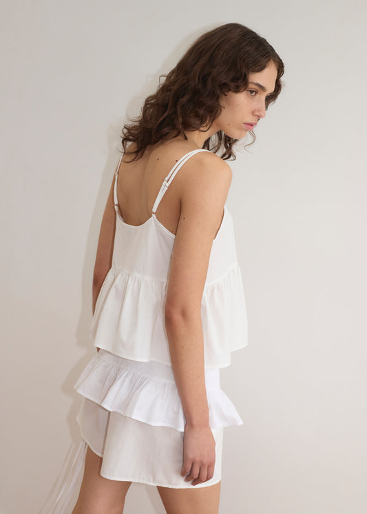 A woman with wavy brown hair wears a sleeveless white ruffled top and a matching skirt paired with the Flounce Belt - White, standing against a plain light-colored wall and looking to the side.