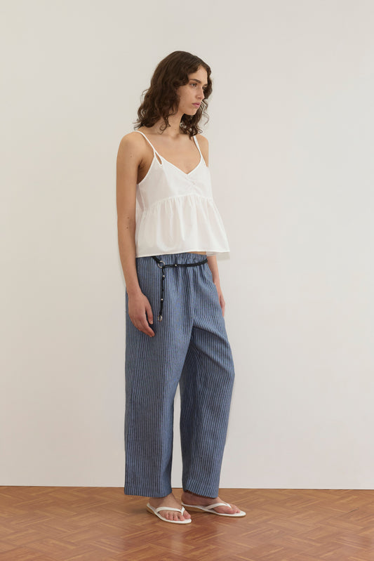 A person with wavy brown hair wears the Bind Top - White from Deiji Studios, blue pinstriped wide-leg pants, and white sandals, standing against a plain light-colored wall on a wooden floor.