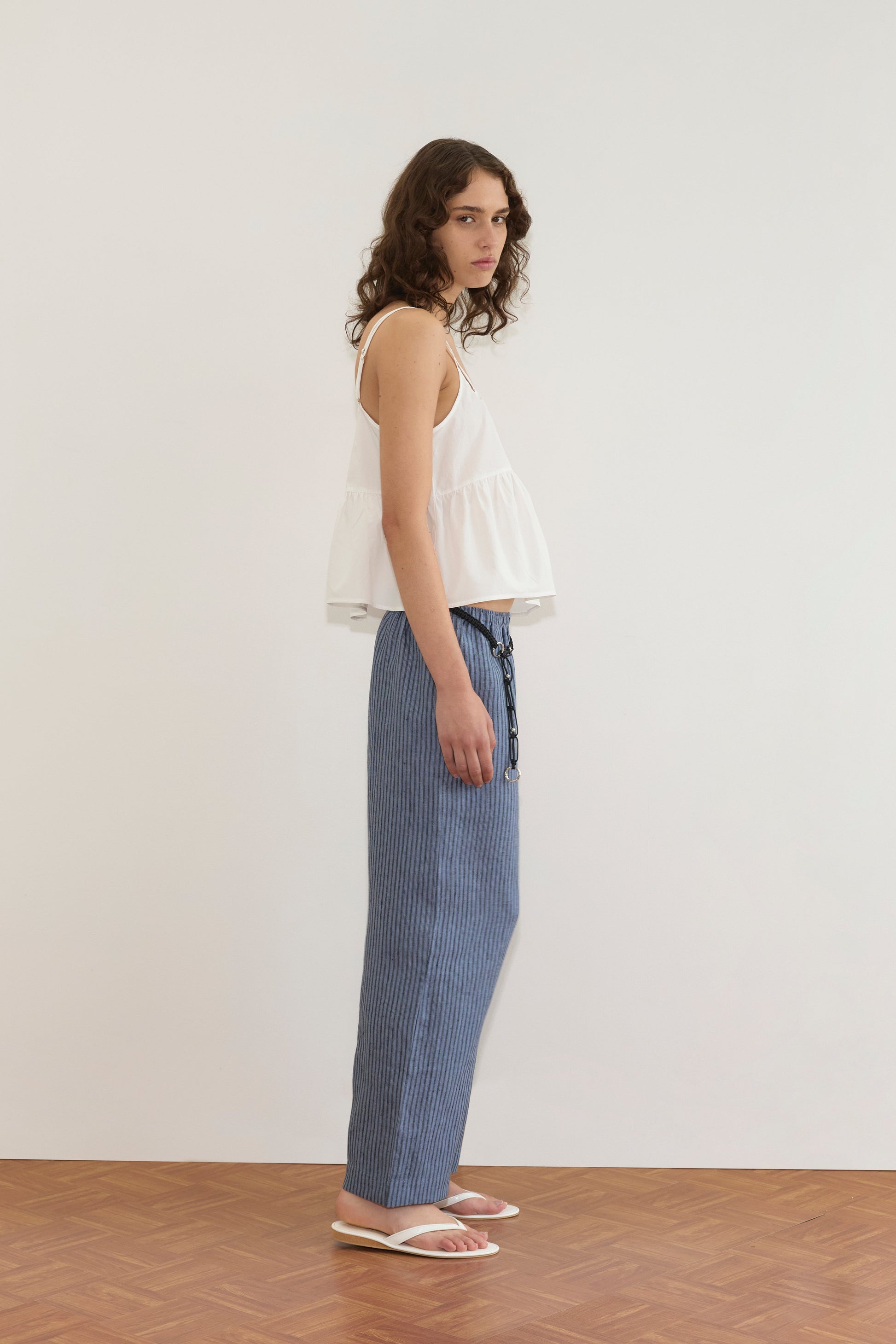 A person with wavy brown hair stands sideways on a wooden floor, wearing a white sleeveless top, Linen Ease Trouser - Azure Stripe, and white sandals, against a plain white wall.