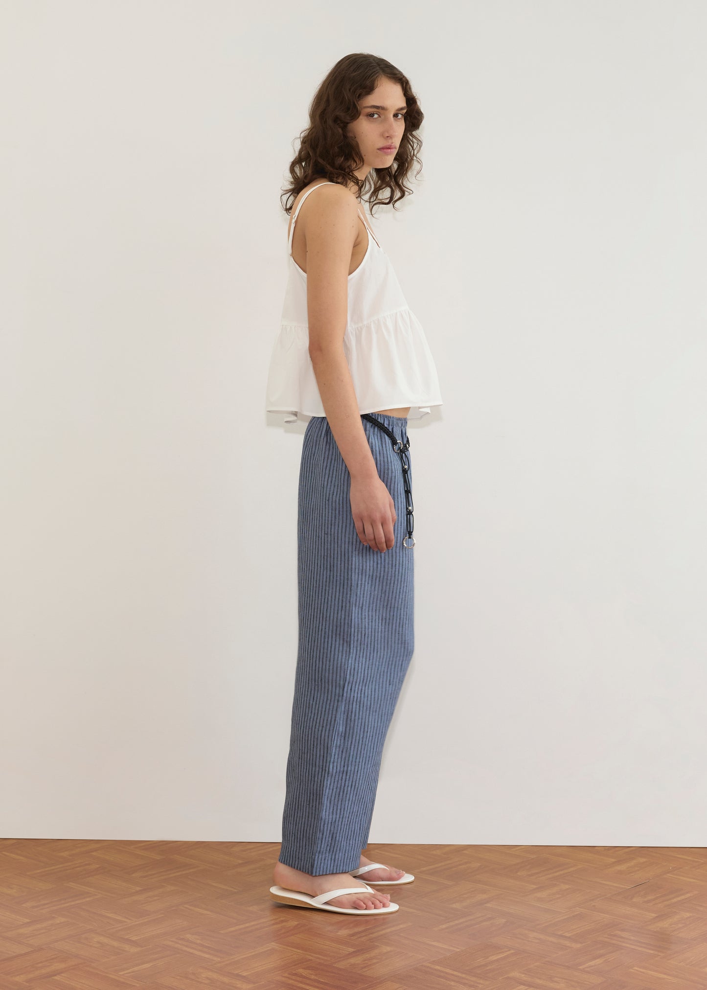 A person with wavy brown hair, wearing the Bind Top - White from Deiji Studios, blue pinstriped pants, and white sandals stands sideways on a wooden floor against a plain white wall.