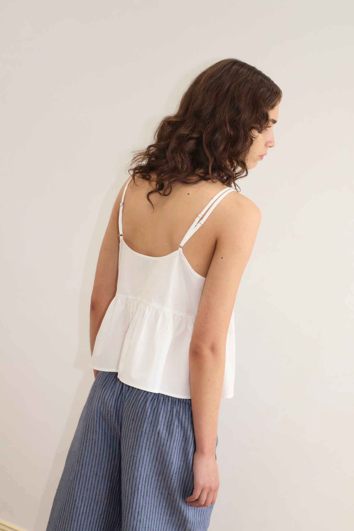 Back view of a woman wearing the Bind Top - White, a cami-style peplum top with double straps from Deiji Studios, paired with blue striped wide-leg trousers.