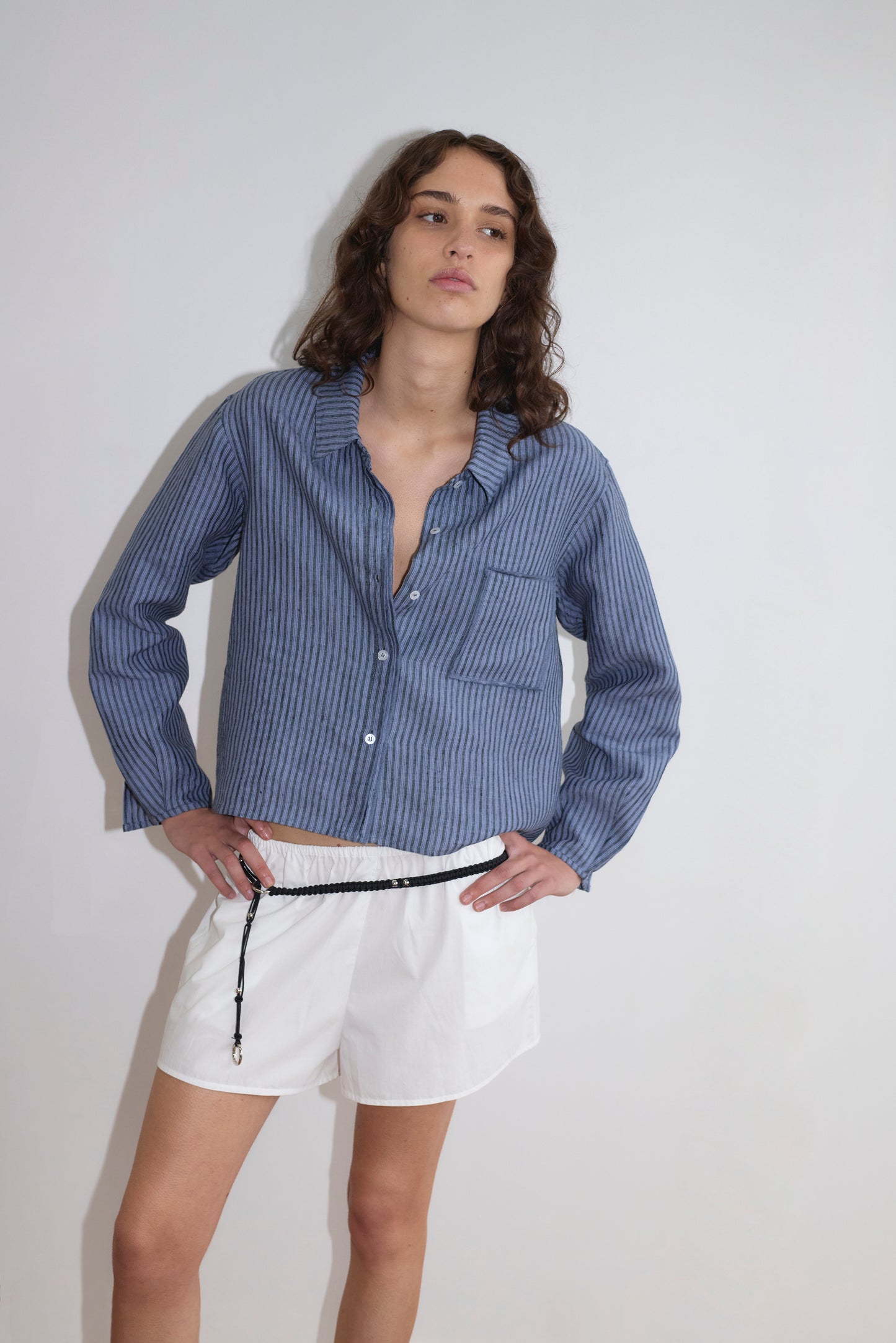 A person with wavy brown hair wears the Deiji Studios Double Placket Shirt - Azure Stripe and white shorts with a black drawstring, standing against a plain white background with one hand on their hip.