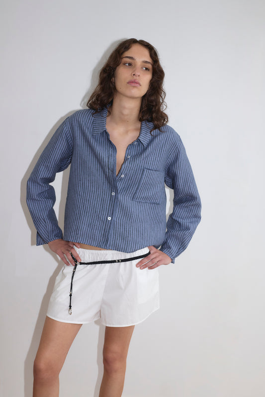 A person with wavy brown hair wears the Deiji Studios Double Placket Shirt - Azure Stripe and white shorts with a black drawstring, standing against a plain white background with one hand on their hip.