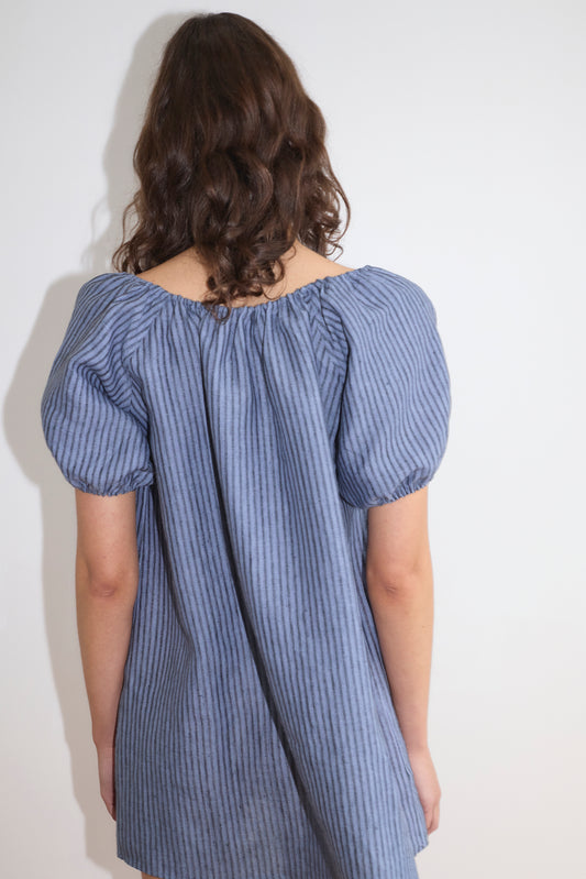 A person with wavy brown hair stands facing away from the camera, wearing Deiji Studios' Smock Dress - Azure Stripe, a loose-fitting blue short-sleeved dress with azure stripes, against a plain white background.