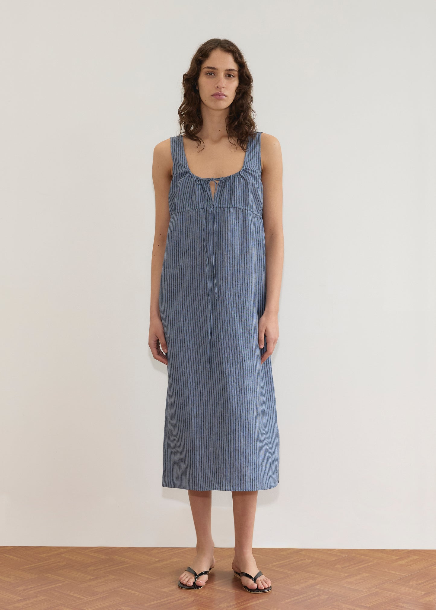 A woman with wavy brown hair wears the Sleeveless Cord Dress - Azure Stripe from Deiji Studios and black sandals. She stands on a wooden floor in front of a plain white wall, looking directly at the camera.