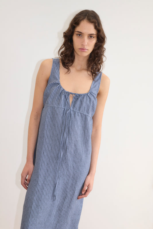 Woman with wavy brown hair stands against a white background, wearing the Deiji Studios Sleeveless Cord Dress - Azure Stripe, featuring blue and white stripes, a scoop neckline, front tie detail, and midi length.