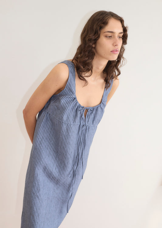 A person with wavy brown hair wears the Sleeveless Cord Dress - Azure Stripe by Deiji Studios, a loose, striped midi dress with a tie neckline, standing against a plain light background and leaning forward with hands behind their back.
