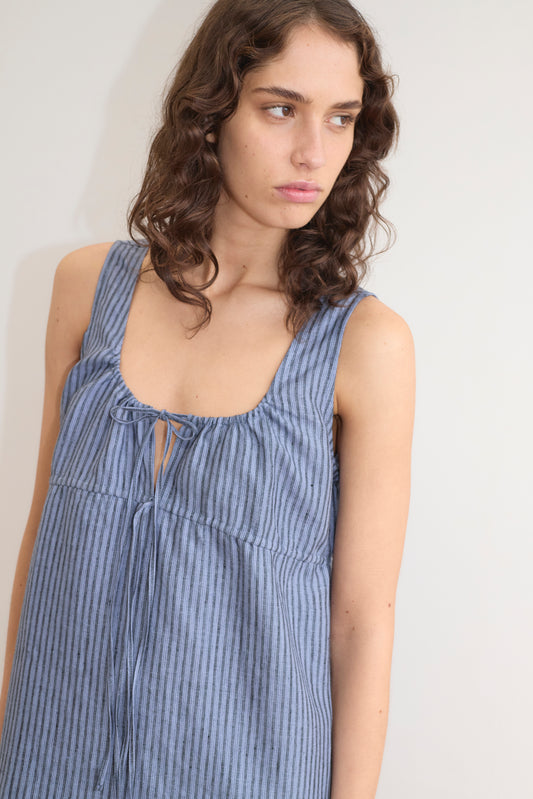 A person with wavy brown hair wearing the Sleeveless Cord Dress - Azure Stripe by Deiji Studios, featuring a front tie, looks to the side against a plain light background.