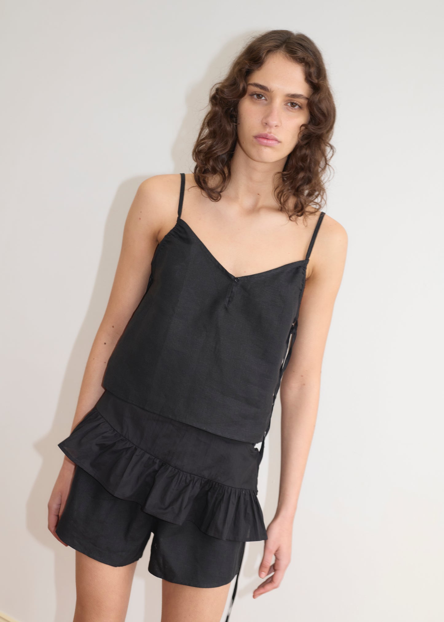 A person with wavy brown hair stands against a white wall, wearing a black sleeveless top and matching Deiji Studios black ruffled shorts styled with the Flounce Belt - Black, looking at the camera with a neutral expression.