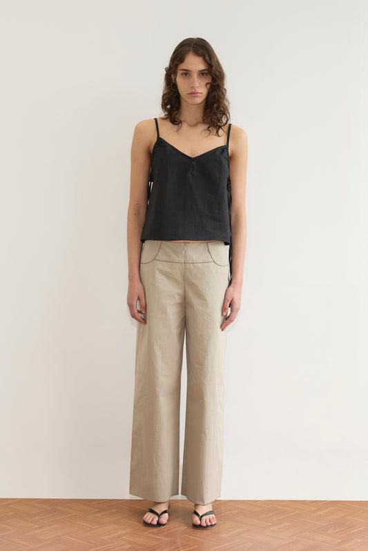 A person with wavy brown hair stands against a plain white wall wearing the Deiji Studios V Neck Cami - Black, beige wide-leg pants, and black sandals on a wooden floor.