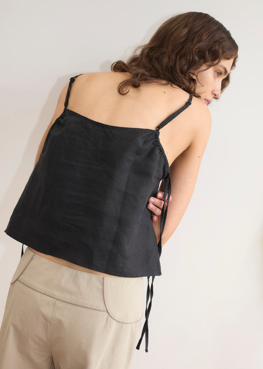 A woman with wavy brown hair, facing away, wears the V Neck Cami - Black by Deiji Studios and beige pants. Her right arm is behind her back against a plain off-white wall.