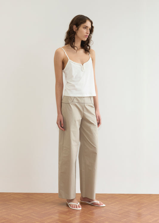 A woman stands indoors in a white camisole, Panelled Pant - Stone with a straight-leg silhouette, and white sandals. She has wavy brown hair and looks slightly to the side against a minimalist background inspired by Deiji Studios.
