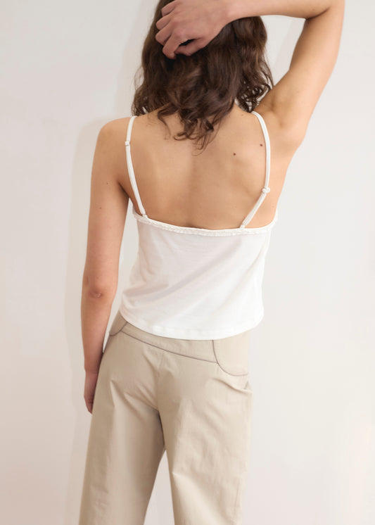 A person with wavy brown hair, facing away from the camera, wears a white camisole and the Panelled Pant - Stone by Deiji Studios in a straight-leg silhouette, with one arm bent and their hand resting on the back of their head.