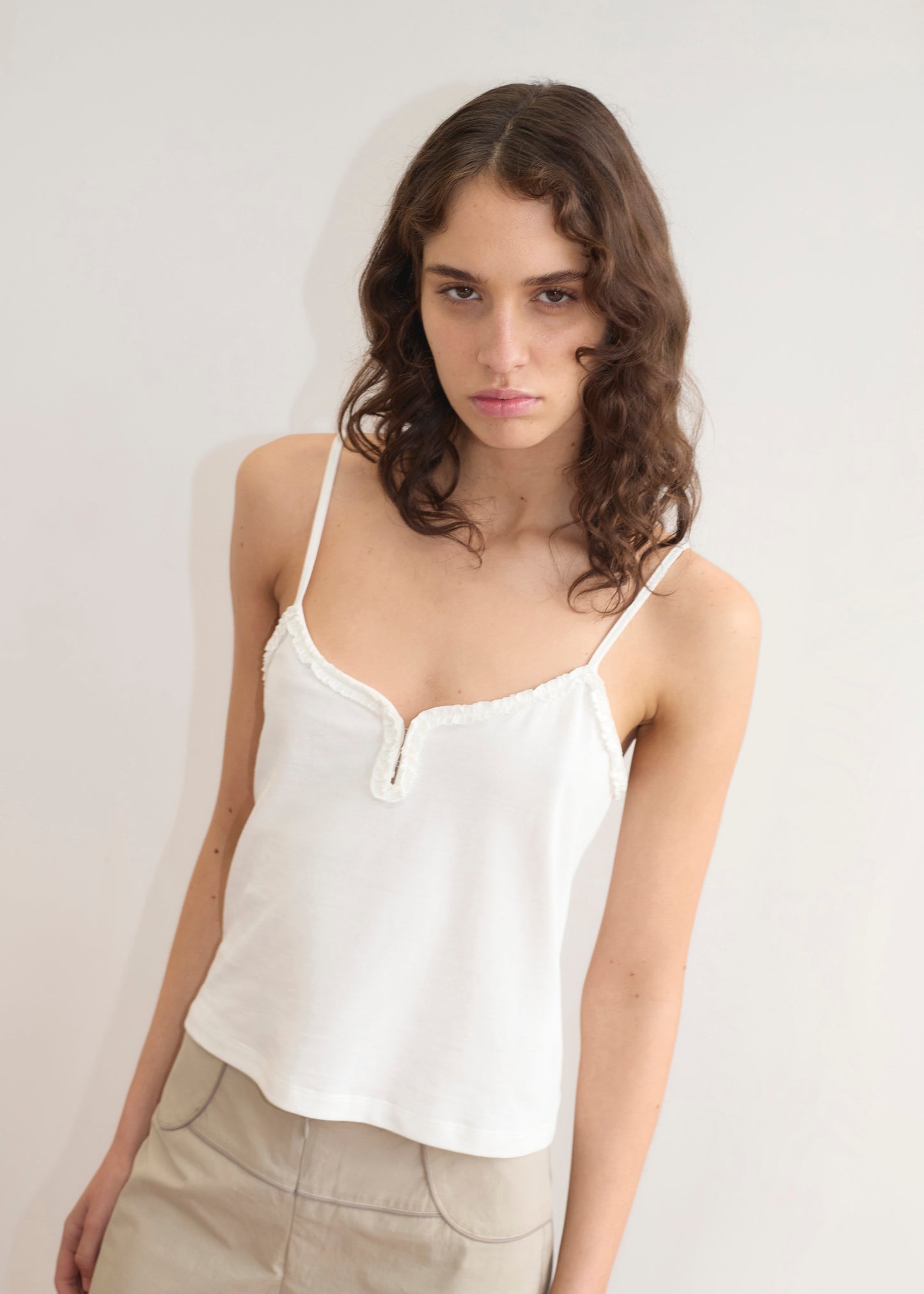 A young woman with wavy brown hair wears the Deiji Studios Clasp Singlet in white and beige pants, standing against a plain light wall, looking slightly toward the camera with a neutral expression.