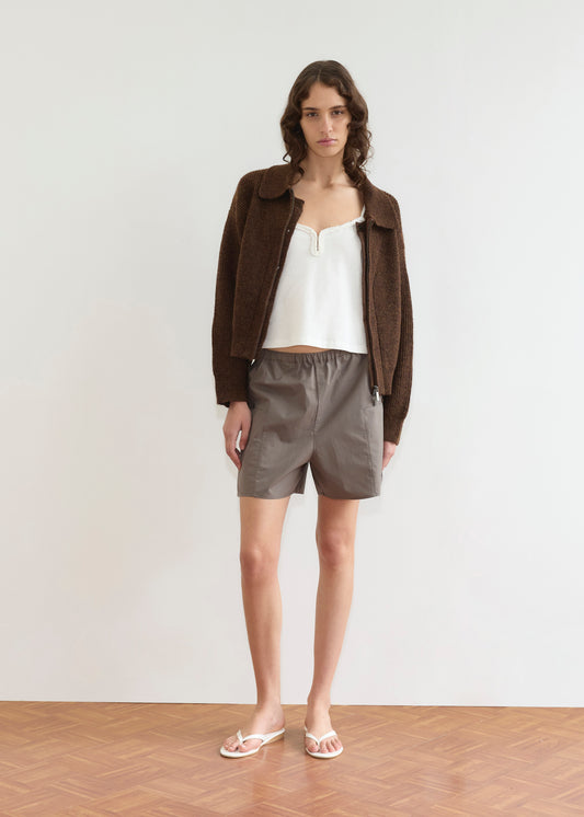 A woman stands indoors on a wooden floor, facing forward against a plain white wall. She wears the Double Layer Cardigan - Brown from Deiji Studios over a white camisole, gray shorts, and white flip-flops.