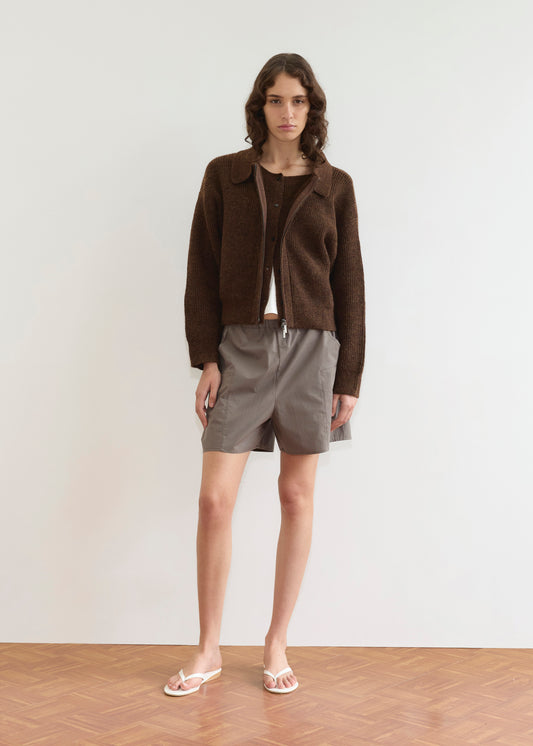 A person with wavy brown hair stands against a plain white wall, wearing a Deiji Studios Double Layer Cardigan - Brown over a matching top, gray drawstring shorts, and white flip-flops on a wooden floor.