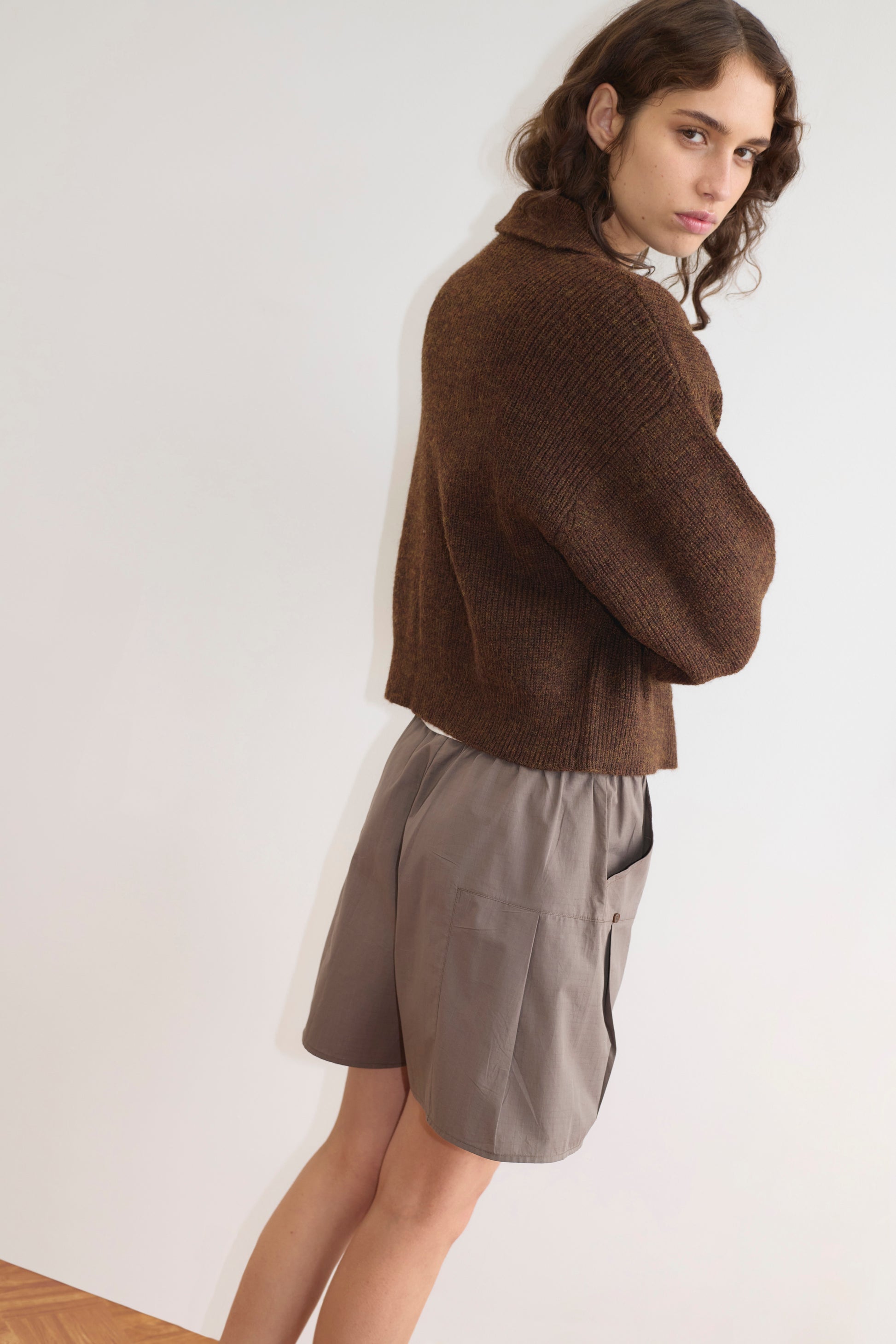 A person with wavy brown hair stands sideways, looking at the camera. They wear a cozy Double Layer Cardigan - Brown by Deiji Studios over loose gray shorts, set against a plain light background.