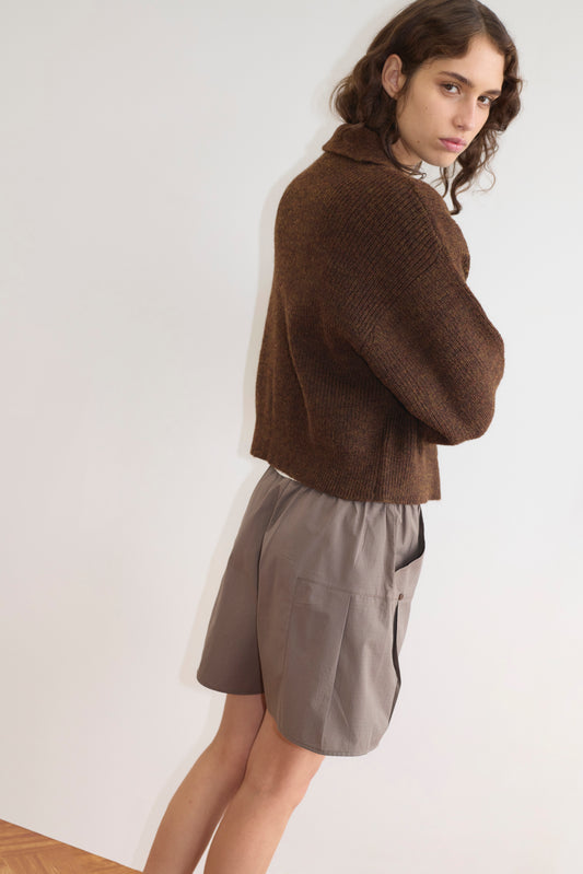 A person with wavy brown hair stands sideways, looking at the camera. They wear a cozy Double Layer Cardigan - Brown by Deiji Studios over loose gray shorts, set against a plain light background.