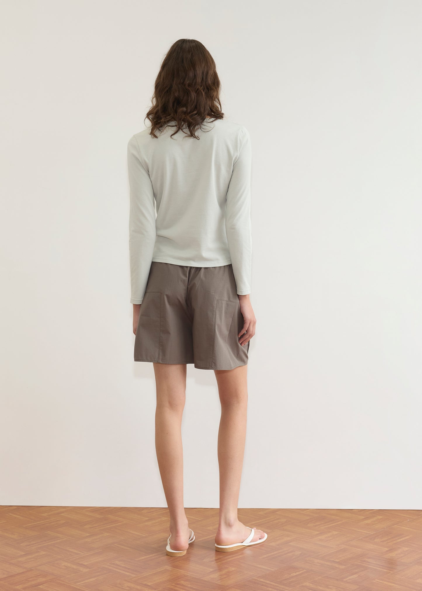 A person with wavy brown hair stands facing a white wall, wearing a Deiji Studios Clasp Longsleeve in Grey Blue, loose brown shorts, and white flip-flops on a wooden floor.