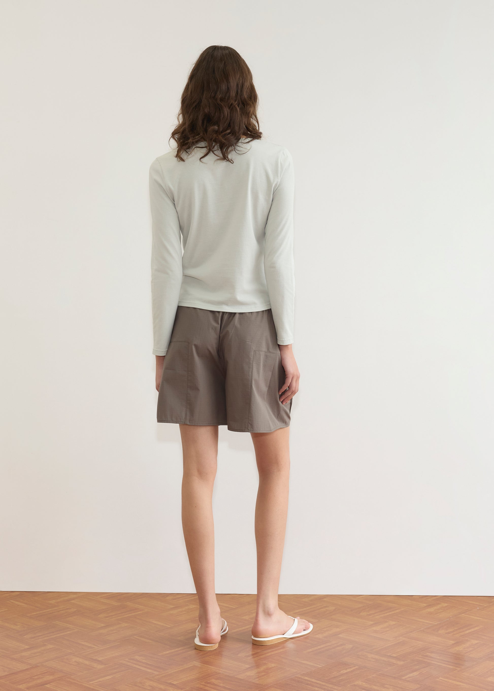 A person with wavy brown hair stands facing a white wall, wearing a Deiji Studios Clasp Longsleeve in Grey Blue, loose brown shorts, and white flip-flops on a wooden floor.