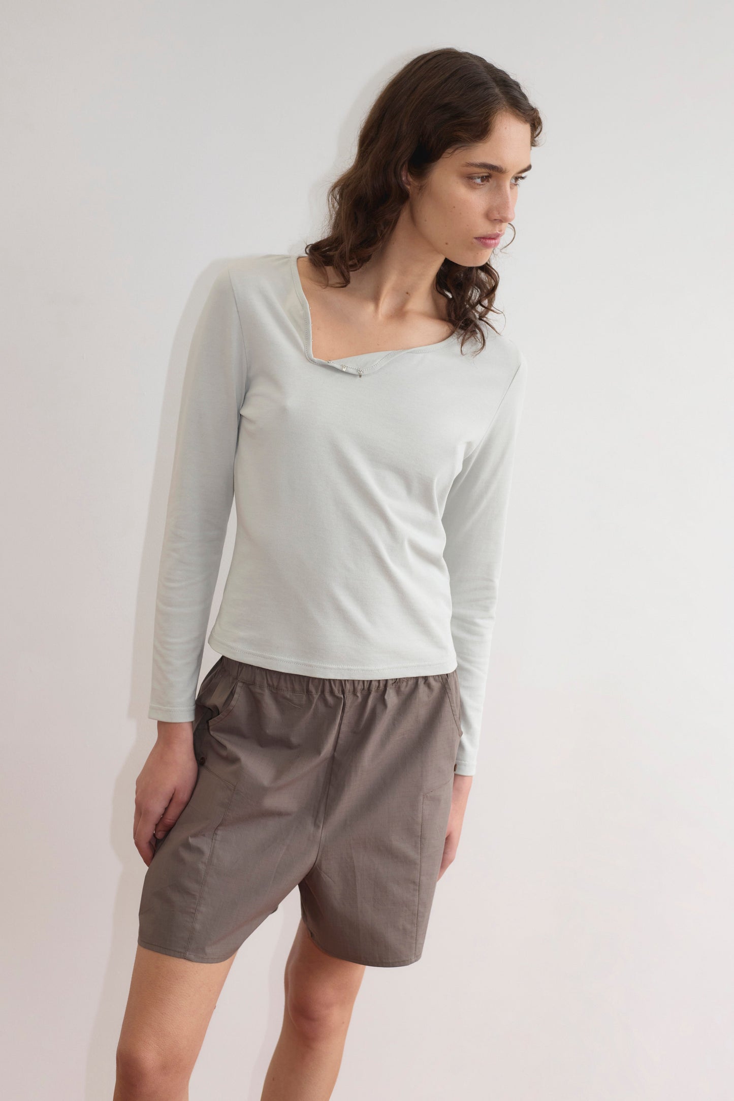 A person with wavy brown hair, wearing the Clasp Longsleeve - Grey Blue by Deiji Studios and loose brown shorts, stands against a plain white background, looking to the side.