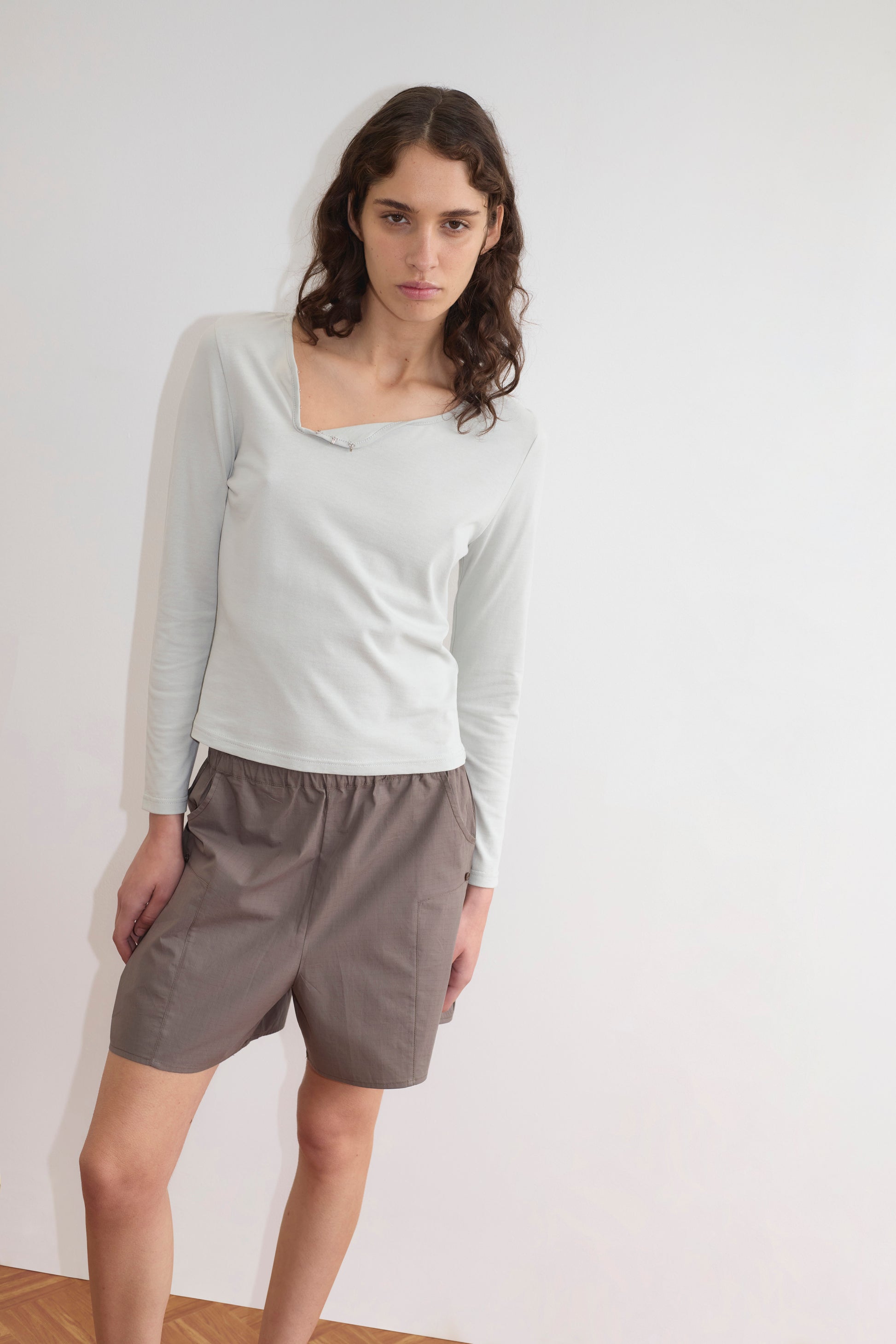 A person with long, wavy hair stands indoors against a plain white wall, wearing a Deiji Studios Clasp Longsleeve in Grey Blue with an asymmetrical neckline and loose taupe shorts.