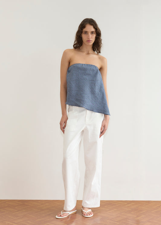 A person stands indoors by a plain white wall, wearing the Line Top - Azure Stripe with a relaxed drape, paired with white wide-leg pants and sandals on a wooden floor for a minimalistic, modern vibe.