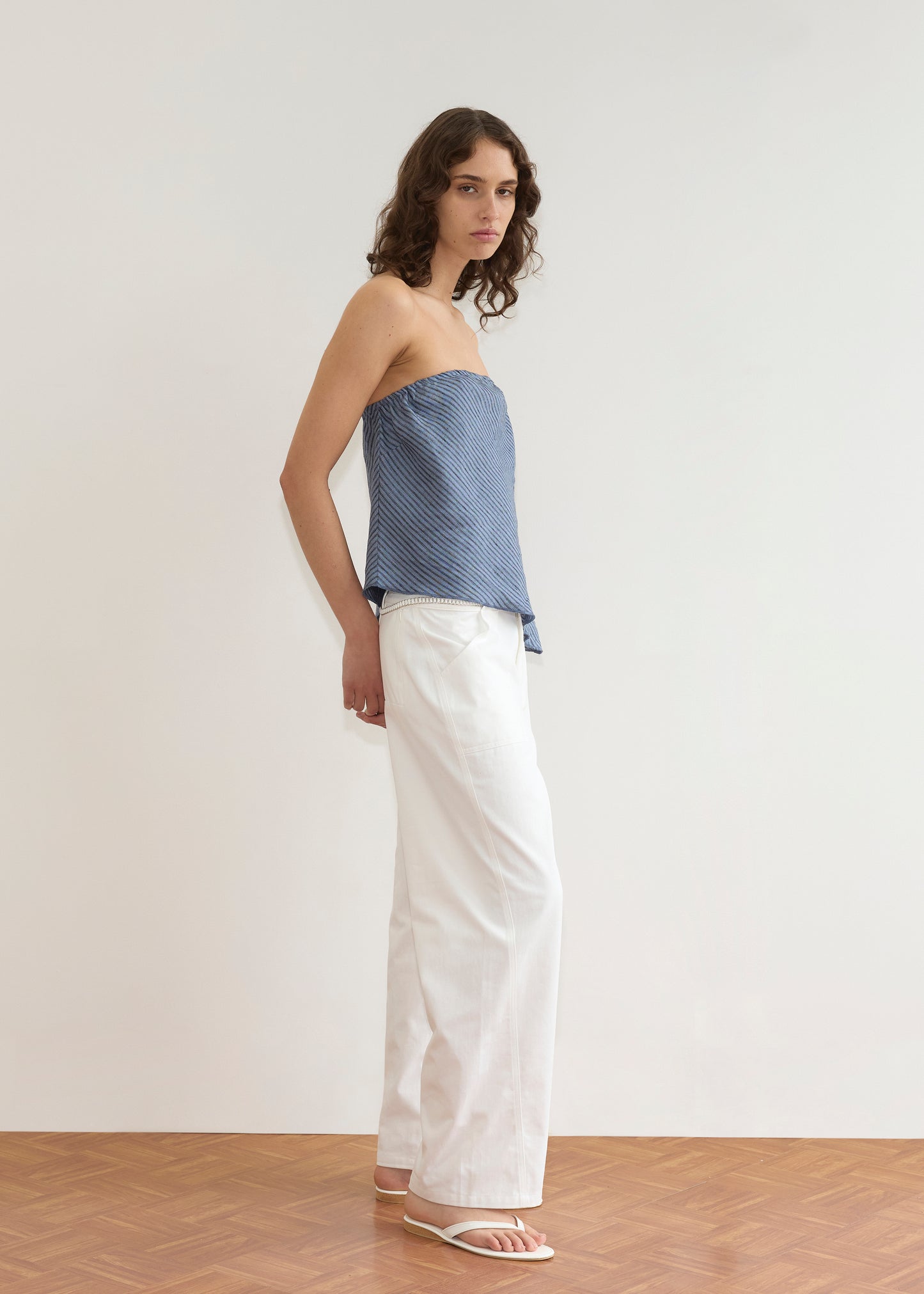 A person with wavy brown hair stands indoors against a plain white wall, wearing the Line Top - Azure Stripe, paired with loose white pants and white sandals, looking at the camera with a neutral expression.