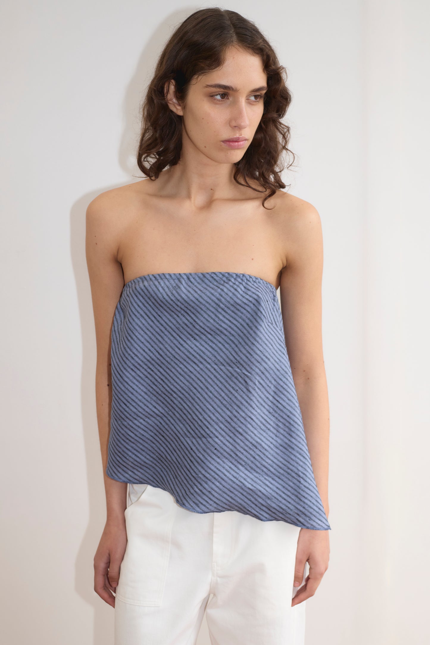 A woman with wavy brown hair models the Line Top - Azure Stripe, a strapless blue top with diagonal stripes and a relaxed drape, paired with white pants against a plain light-colored wall.