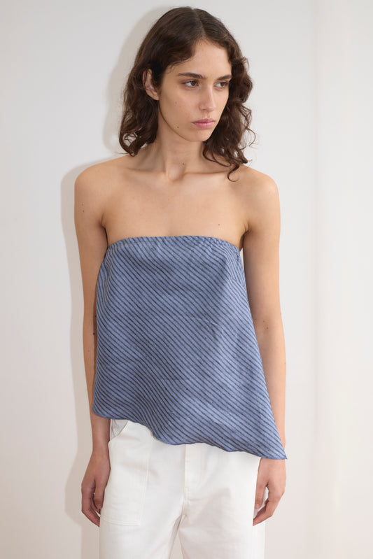 A woman with wavy brown hair models the Line Top - Azure Stripe, a strapless blue top with diagonal stripes and a relaxed drape, paired with white pants against a plain light-colored wall.