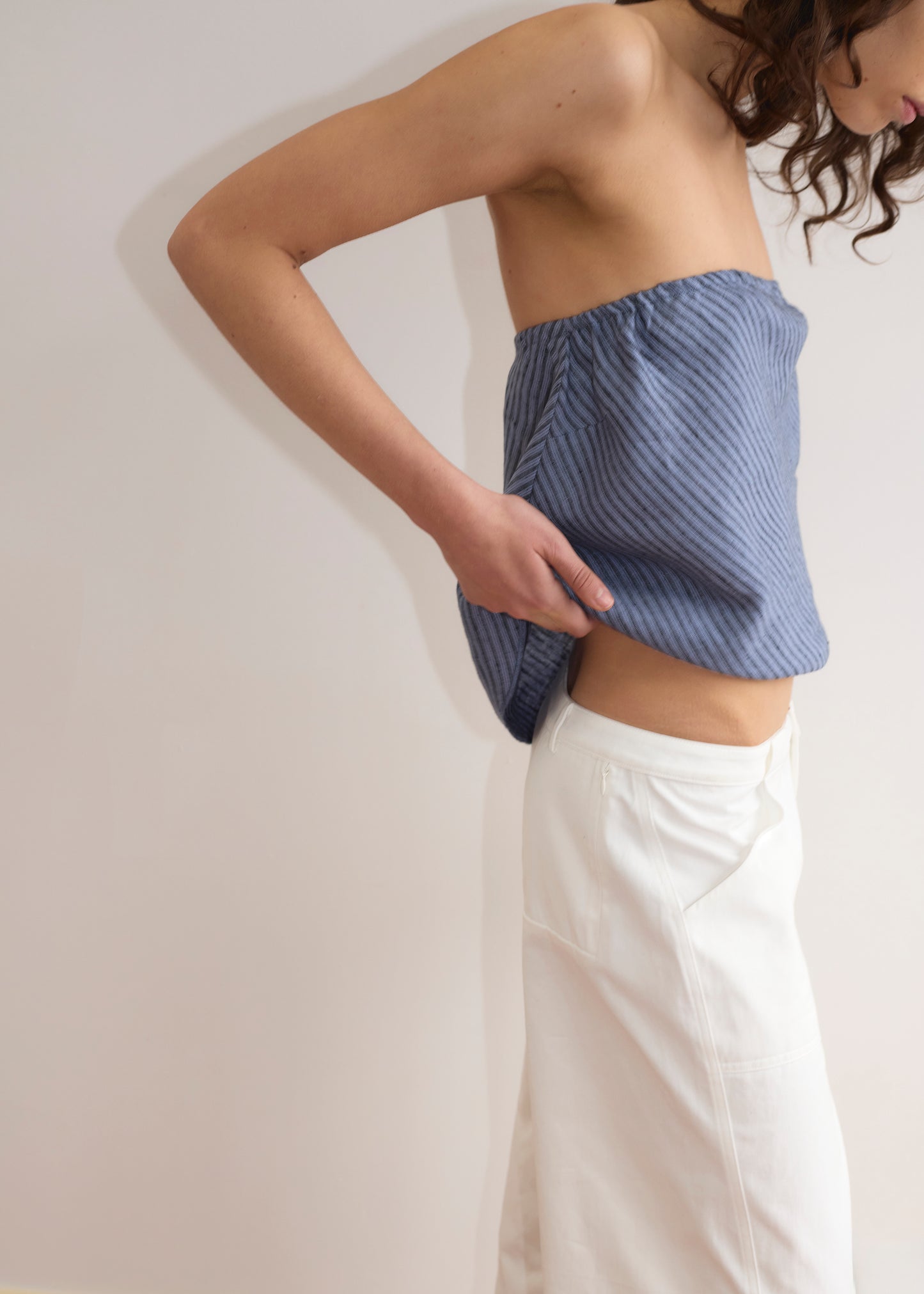 A person wearing the Line Top - Azure Stripe and white pants stands sideways against a plain background, adjusting their pants with one hand. Their blue lightweight strapless top features subtle stripes; their face is partially out of frame.