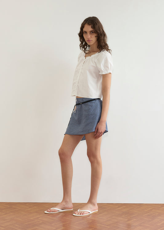 A young woman with wavy brown hair stands indoors on a wooden floor, wearing the Close Shirt in White, a blue striped mini skirt, and white sandals. She looks at the camera against a plain white wall.