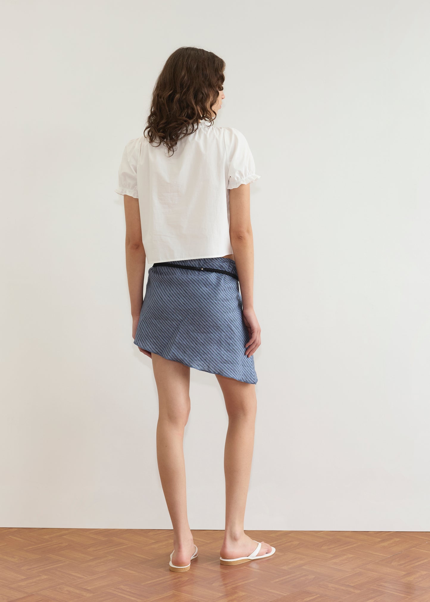 A woman with wavy brown hair stands facing away, wearing the Close Shirt - White, a blue and white striped wrap skirt, and white sandals on a wooden floor against a plain white wall.