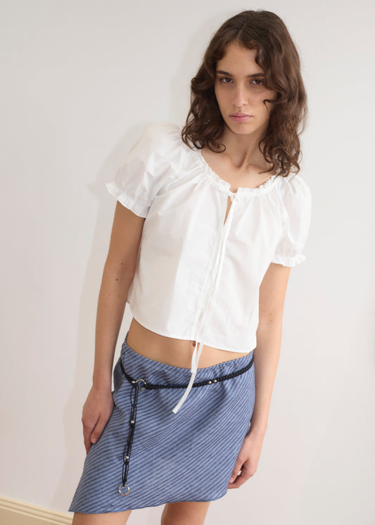 A woman with wavy brown hair wears the Line Top - Azure Stripe, a white lightweight top with a neckline tie, paired with a blue striped skirt and black belt, standing against a light-colored wall.