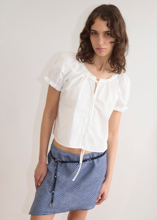 A young woman with wavy brown hair models the Close Shirt - White, a cropped relaxed short sleeve top with a tie neckline, paired with a blue diagonally striped mini skirt and black cord belt, standing against a plain white background.