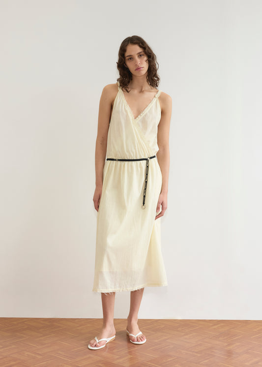 A woman with wavy brown hair stands on a wooden floor, wearing the Crossed Dress - Soft Yellow Stripe with a thin black belt and white sandals, against a plain white background.