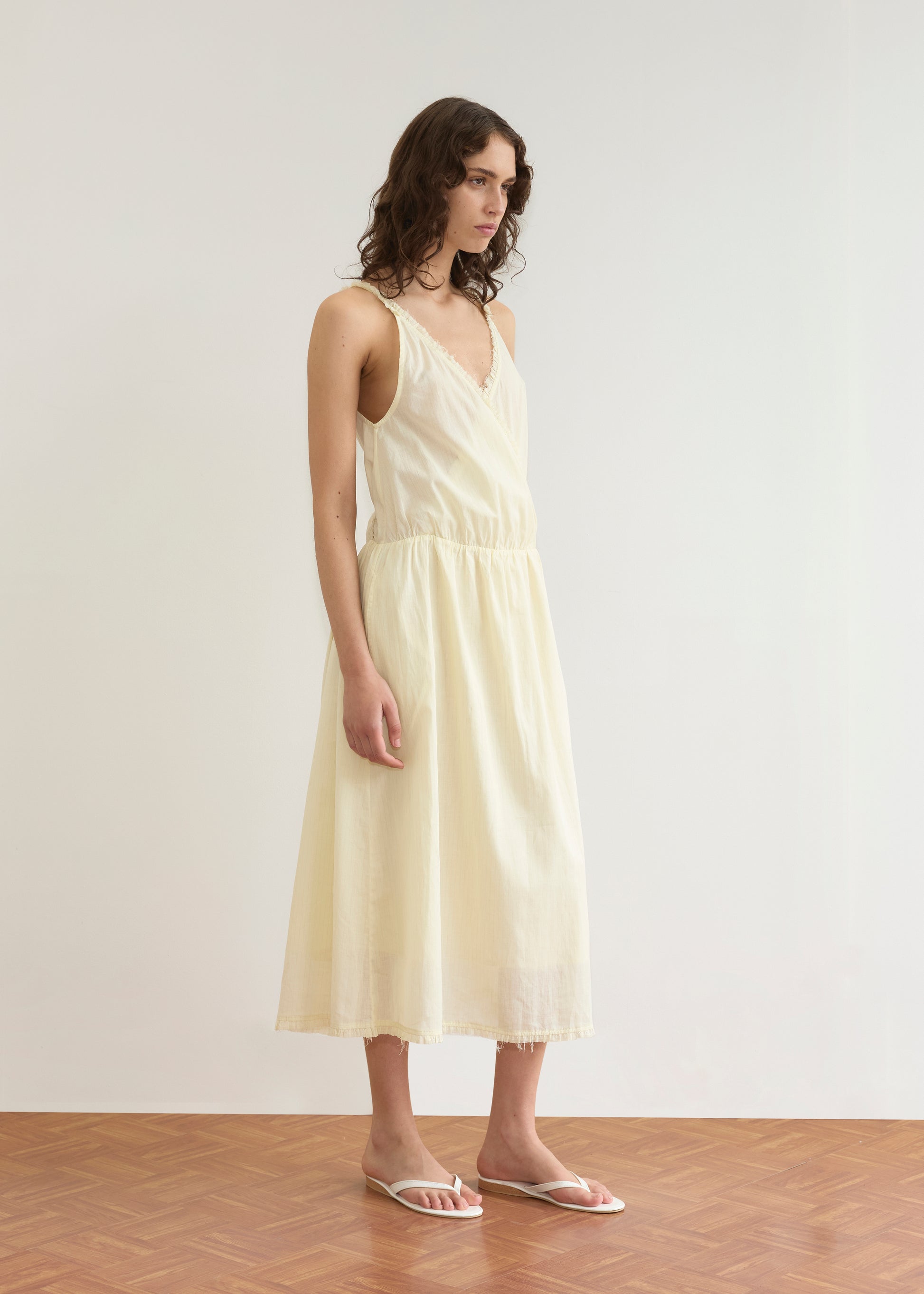 A person with wavy brown hair wears the Crossed Dress - Soft Yellow Stripe by Deiji Studios, featuring a crossover v-neckline and white sandals, standing on a wooden floor against a plain white wall, gazing slightly to the side.