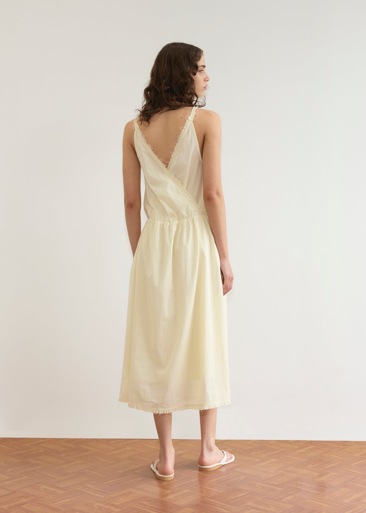 A woman with wavy brown hair stands on a wooden floor, facing away in the Crossed Dress - Soft Yellow Stripe by Deiji Studios—a sleeveless midi dress with a deep V-back—paired with white sandals, set against a plain white background.