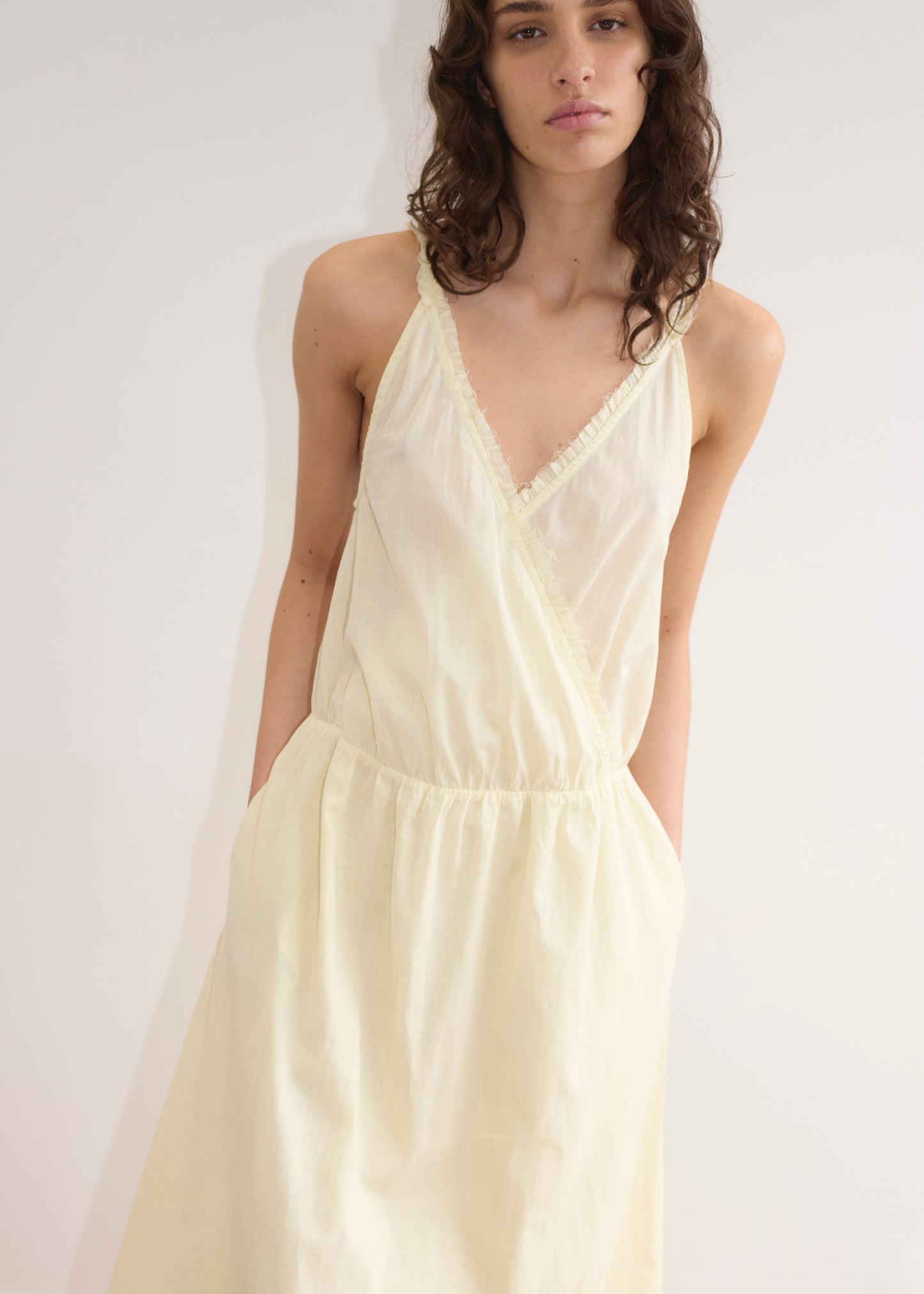 A woman with wavy brown hair wears the Crossed Dress - Soft Yellow Stripe by Deiji Studios, featuring a crossover v-neckline and pockets, standing against a plain white background.