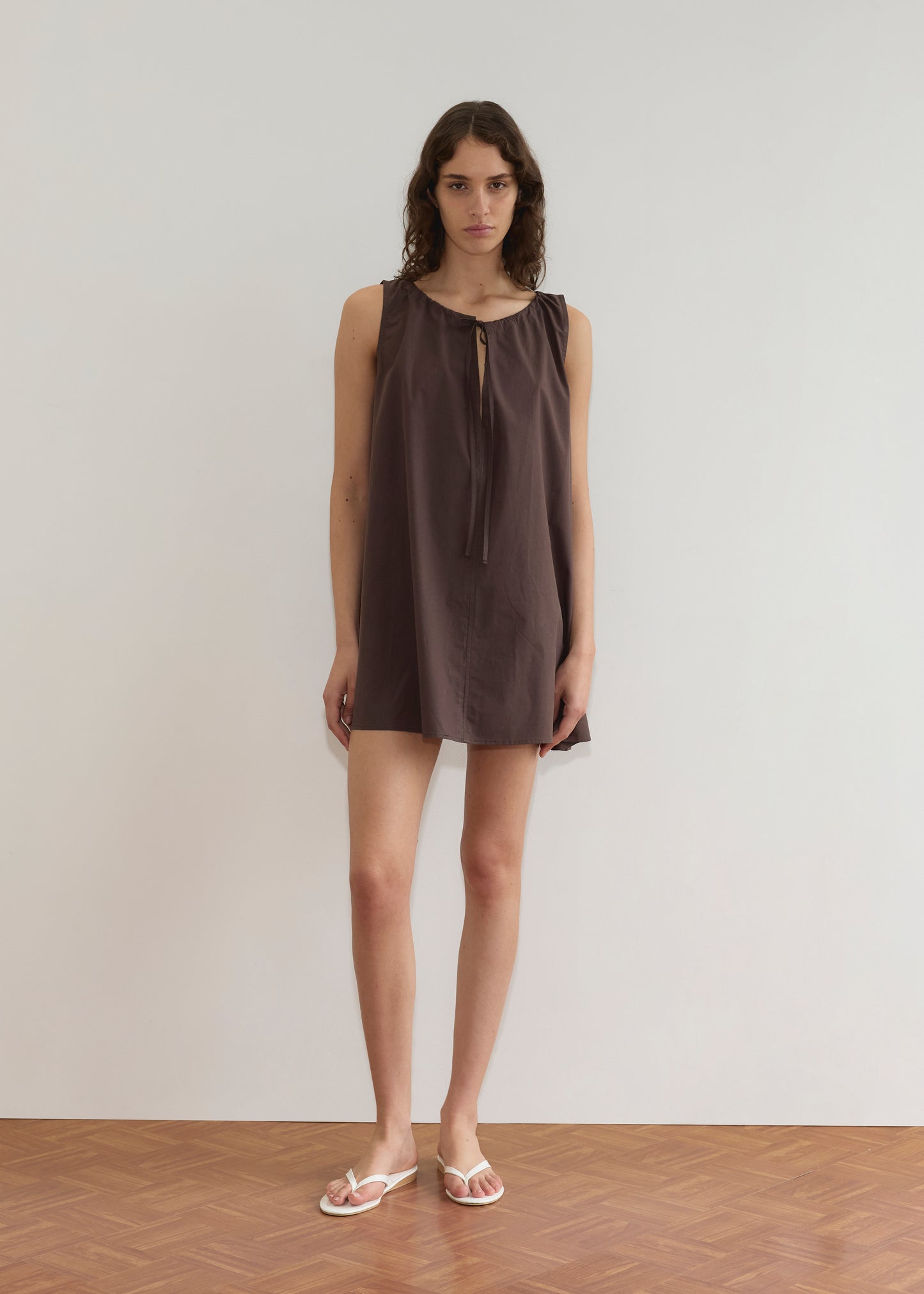 A person with wavy brown hair stands against a plain white wall, wearing the Drawcord Dress - Sepia by Deiji Studios with an a-line silhouette, sleeveless and loose-fitting, paired with white sandals on a wooden floor.