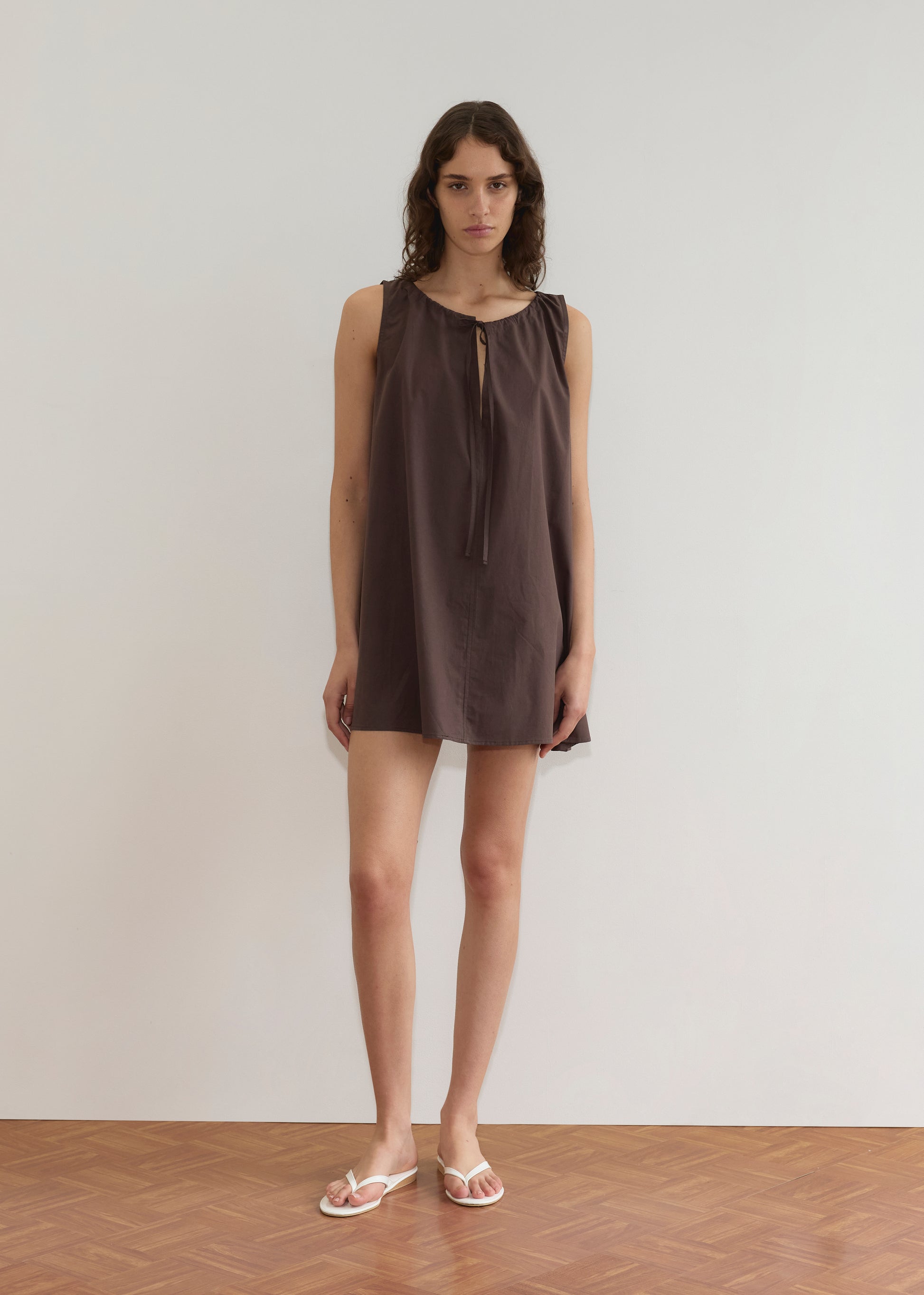 A person with wavy brown hair stands against a plain white wall, wearing the Drawcord Dress - Sepia by Deiji Studios with an a-line silhouette, sleeveless and loose-fitting, paired with white sandals on a wooden floor.