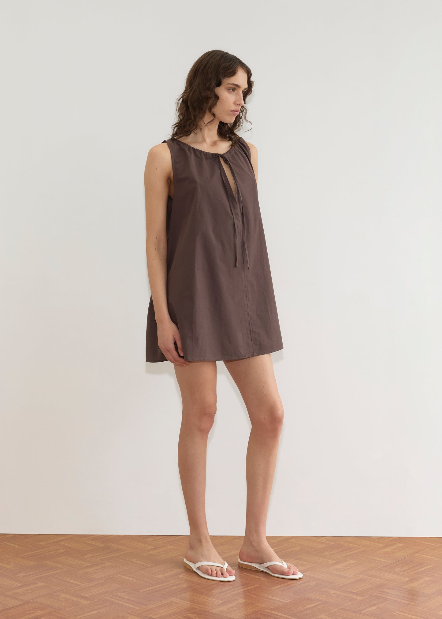 A woman stands indoors in the Deiji Studios Drawcord Dress - Sepia, a loose brown mini with an A-line silhouette and adjustable neckline tie. She pairs it with white sandals, her wavy hair framing her face as she looks to the side against a white wall.