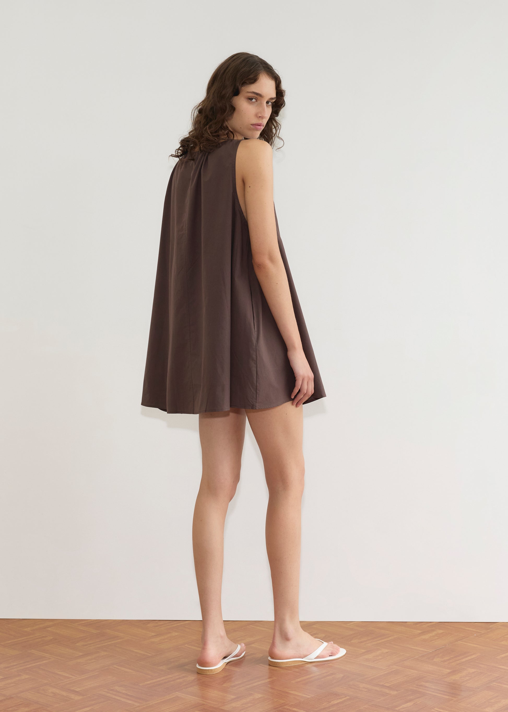 A person with long, wavy brown hair stands sideways on a wooden floor, wearing the Deiji Studios Drawcord Dress - Sepia with an a-line silhouette and white flip-flops, posed against a plain white wall.