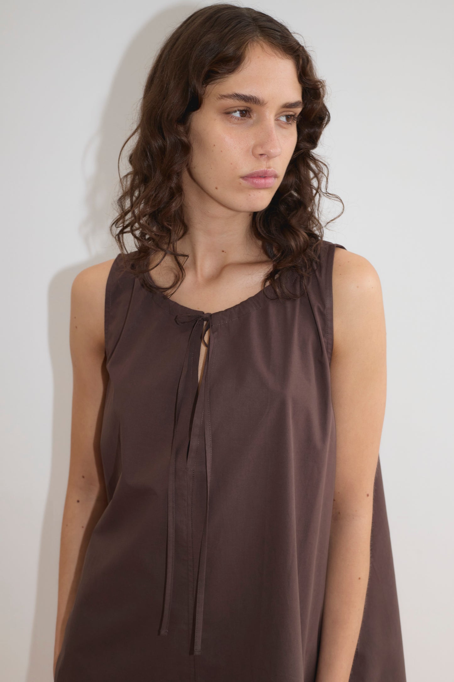 A woman with wavy brown hair wears the Drawcord Dress - Sepia by Deiji Studios, a sleeveless, loose-fit brown dress with tie neckline, standing against a plain white background and looking to her left.