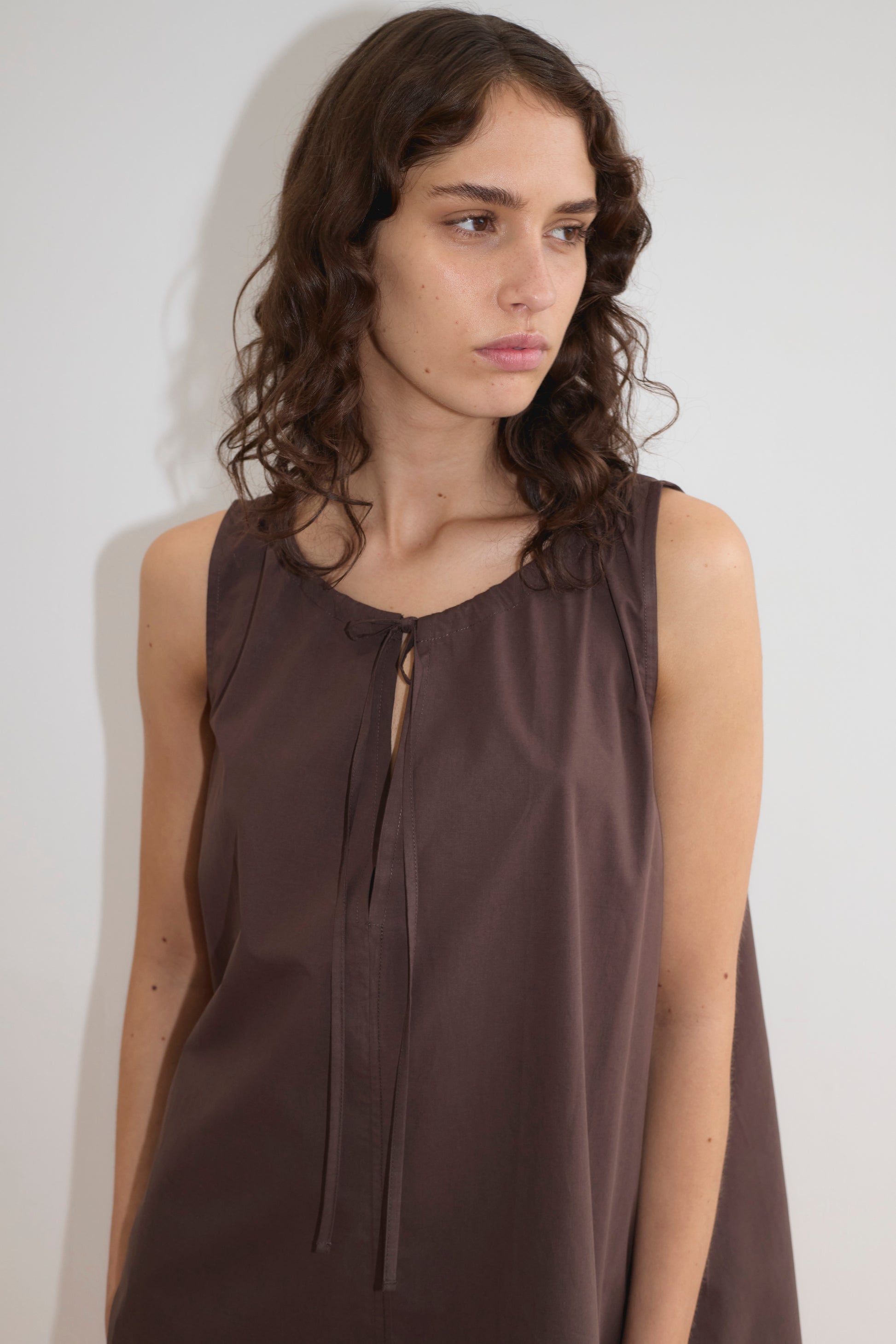 A woman with wavy brown hair wears the Drawcord Dress - Sepia by Deiji Studios, a sleeveless, loose-fit brown dress with tie neckline, standing against a plain white background and looking to her left.